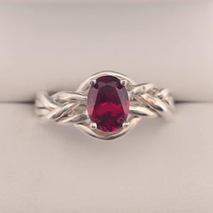 Op de afbeelding: Een zilveren ring met een gevlochten band en een rode ovale edelsteen.
