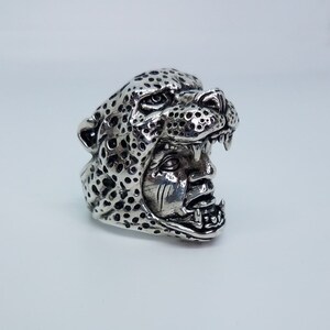 May include: A silver ring with a detailed design of a leopard's head with a human face inside. The ring is made of metal and has a textured surface.
