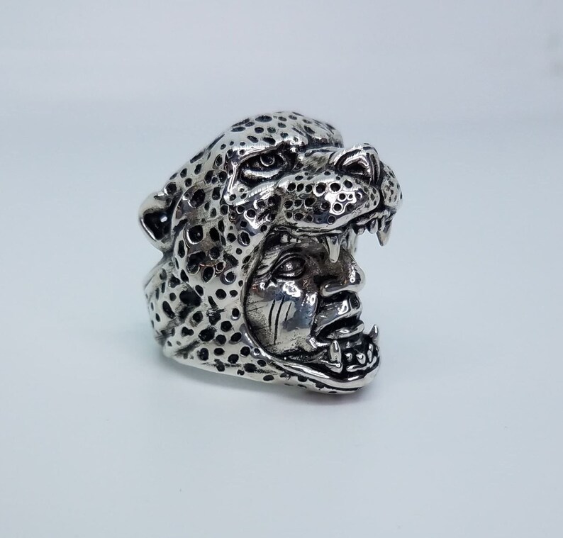 May include: A silver ring with a detailed design of a leopard's head with a human face inside. The ring is made of metal and has a textured surface.