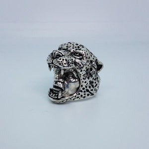 May include: A silver ring with a detailed cheetah head design. The cheetah's mouth is open, showing its teeth. The ring is a statement piece with a unique and bold design.