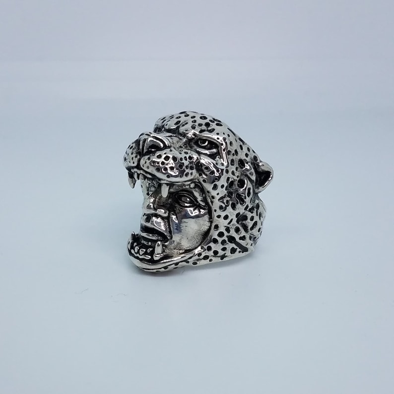 May include: A silver ring with a detailed cheetah head design. The cheetah's mouth is open, showing its teeth. The ring is a statement piece with a unique and bold design.