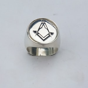K&ouml;nnte beinhalten: Ein silberner Ring mit einer runden, gravierten Oberseite, die ein freimaurerisches Symbol zeigt. Das Symbol ist ein Zirkel und ein Winkelma&szlig;, mit einer Wasserwaage oben.