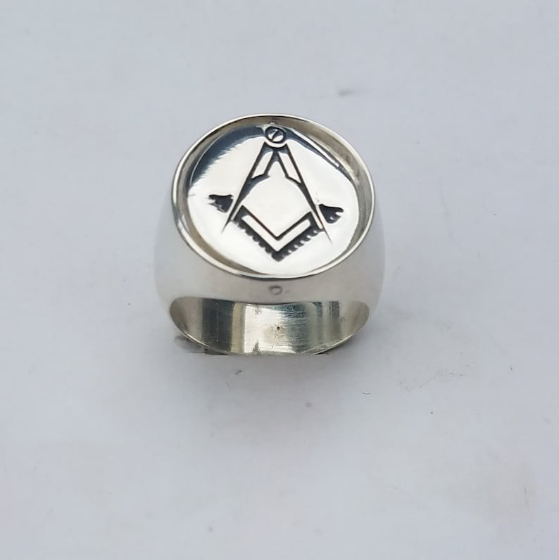 K&ouml;nnte beinhalten: Ein silberner Ring mit einer runden, gravierten Oberseite, die ein freimaurerisches Symbol zeigt. Das Symbol ist ein Zirkel und ein Winkelma&szlig;, mit einer Wasserwaage oben.