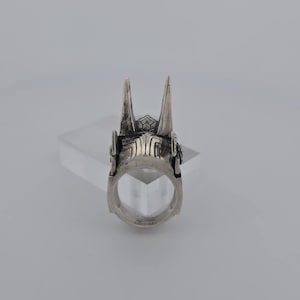 May include: A silver ring with a detailed design featuring two pointed spikes on top. The ring is made of metal and has a smooth, polished finish.