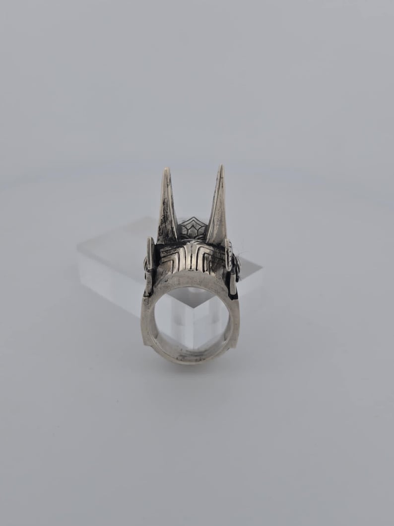 May include: A silver ring with a detailed design featuring two pointed spikes on top. The ring is made of metal and has a smooth, polished finish.