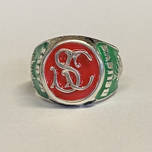 May include: A silver ring with a red enamel center featuring the letters "SC" in a white script. The ring is surrounded by a green enamel band with the word "Believe" inscribed on it.