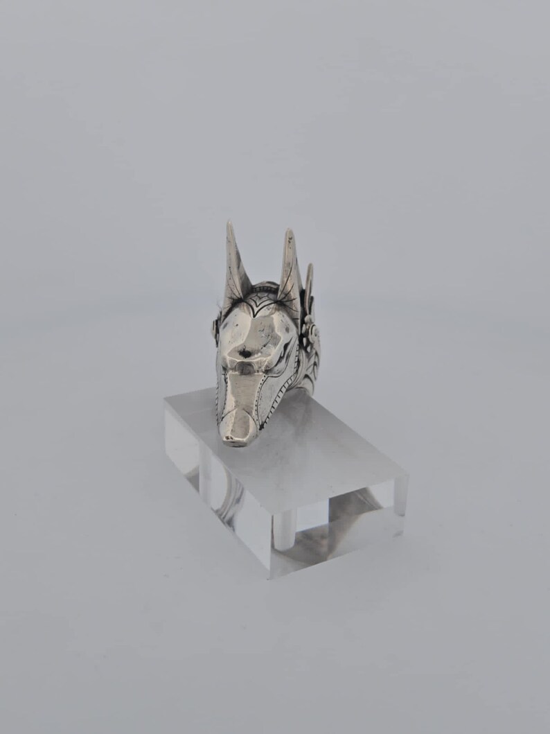 May include: Silver ring with a detailed Anubis head design. The ring is on a clear display stand.
