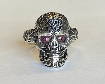 May include: A silver skull ring with intricate detailing and red gemstone eyes. The skull design features ornate patterns on the head and around the jawline. The ring is a bold statement piece.
