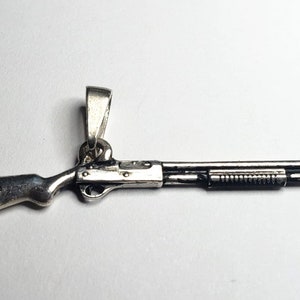 Könnte beinhalten: Silberner Schrotflinten-Anhänger. Der Anhänger ist detailliert mit einem strukturierten Lauf und einer silbernen Öse zum Anbringen an einer Kette.