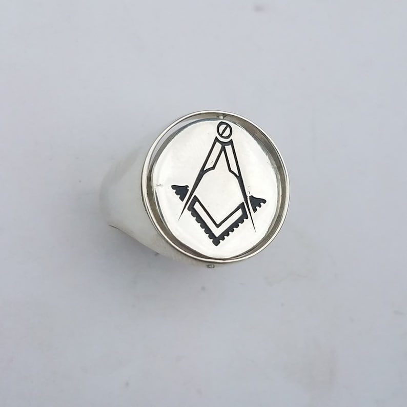 K&ouml;nnte beinhalten: Ein silberner Ring mit einer runden Oberfl&auml;che, auf der ein schwarz-wei&szlig;es, eingraviertes freimaurerisches Symbol, ein Zirkel und ein Winkelma&szlig;, abgebildet sind.