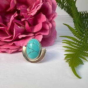 May include: A silver ring with a large, oval turquoise stone. The stone has a dark, web-like pattern. The ring's band has a half-moon design. The ring is set against a backdrop of a pink rose and green fern.