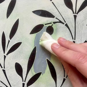 May include: A white stencil featuring a repeating olive branch pattern with cut-out leaves and olives. The stencil is being used with a cloth to apply light green and gray paint. The text LAVABLE / REUTILIZABLE is printed at the bottom.