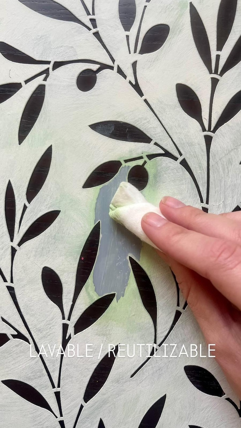 May include: A white stencil featuring a repeating olive branch pattern with cut-out leaves and olives. The stencil is being used with a cloth to apply light green and gray paint. The text LAVABLE / REUTILIZABLE is printed at the bottom.
