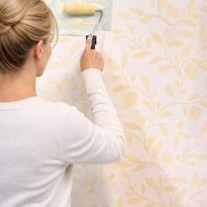 Puede incluir: Una persona aplicando papel pintado con un rodillo. El papel pintado presenta un patrón repetido de hojas y ramas amarillas sobre un fondo blanco. La persona lleva un jersey blanco.