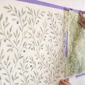 May include: A wall stencil featuring a repeating olive branch pattern with sage green leaves and small dark berries. The stencil is partially applied to a white wall, secured with purple painter's tape, showing the transition from the stencil sheet to the painted wall design.