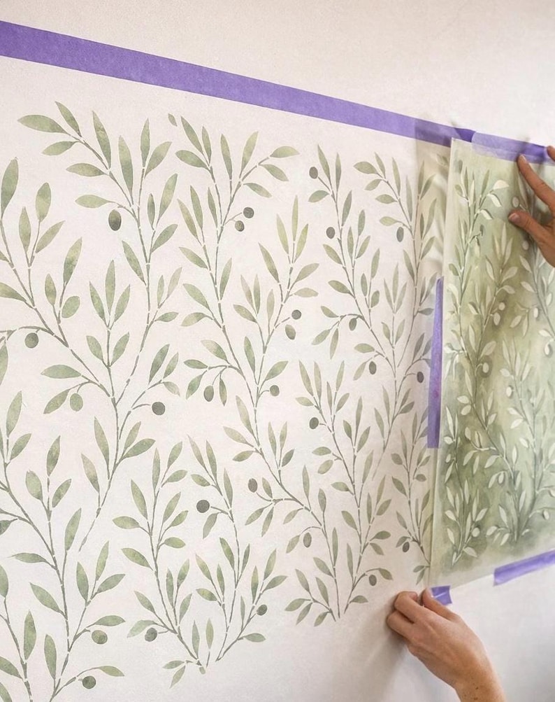 May include: A wall stencil featuring a repeating olive branch pattern with sage green leaves and small dark berries. The stencil is partially applied to a white wall, secured with purple painter's tape, showing the transition from the stencil sheet to the painted wall design.