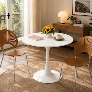 May include: A white round pedestal table with a central column base, accompanied by two chairs featuring light brown wooden seats and backrests with vertical slats, supported by thin silver metal legs, arranged on a round woven rug.