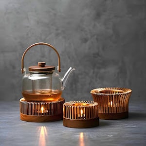 May include: A glass teapot with a wooden handle and lid sits atop a wooden warmer. Two wooden candle holders with lit candles are also present. The items are arranged on a gray surface against a gray background.