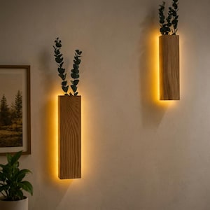 May include: Two rectangular wooden wall sconces with a light wood grain finish, each featuring a hollow top holding green eucalyptus stems. The fixtures emit a warm, soft glow from behind the wood, casting light onto the wall surface.