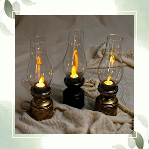 May include: Three decorative oil lamps with clear glass chimneys and flickering LED candle lights. The bases are a mix of black and antique gold, with a handle on one. The lamps are arranged on a neutral-toned fabric.