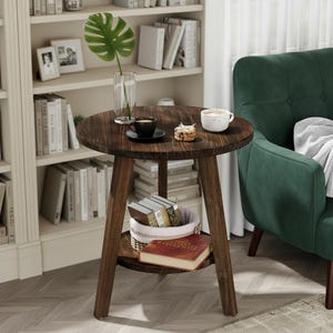 May include: A round dark brown wooden side table with three angled legs and a lower circular shelf. The tabletop holds a clear glass vase with a green leaf, a black coffee cup on a saucer, and a small white bowl. A basket and book sit below.