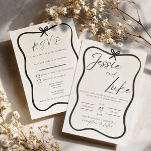 May include: Two cream wedding invitation cards with black text and a black border. One card says "RSVP" and the other says "Jessie and Luke". Both cards have a bow design at the top and are decorated with white flowers.