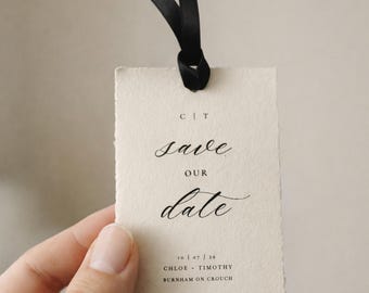 May include: A cream-colored tag with the words "save our date" in elegant script, tied with a black ribbon. The tag also includes the date, names, and location. A simple and stylish wedding announcement.