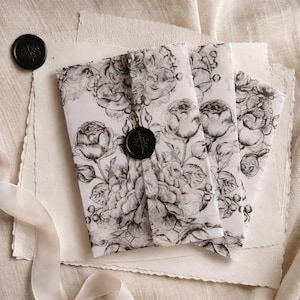 May include: Three folded packages wrapped in white paper with a black and white floral design. Each package is sealed with a black wax seal. The packages are on top of textured paper and a cream-colored fabric.