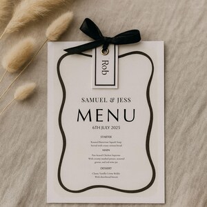 May include: A wedding menu card with a black ribbon and a tag that says "Rob". The menu is for Samuel & Jess, dated July 6th, 2025. The menu includes a starter, main course, and dessert.