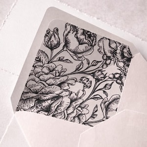 Op de afbeelding: Witte envelop met een bloemenvoering. De voering heeft een gedetailleerde zwart-wit illustratie van rozen en andere bloemen. De envelop is open en onthult het bloemmotief binnenin. Het papier heeft een getextureerd uiterlijk.
