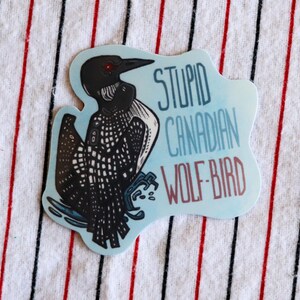 Peut inclure: Un autocollant représentant un huard avec le texte "STUPID CANADIAN WOLF-BIRD". L'oiseau est noir et blanc avec un œil rouge, sur un fond bleu clair.