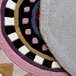 K&ouml;nnte beinhalten: Ein farbenfroher, runder Teppich mit geometrischem Design. Der Teppich hat konzentrische Farbstreifen, darunter Rosa, Lila, Schwarz, Gold und Wei&szlig;. Die R&uuml;ckseite ist ein grauer Stoff. Das Design umfasst Quadrate, Dreiecke und Punkte.