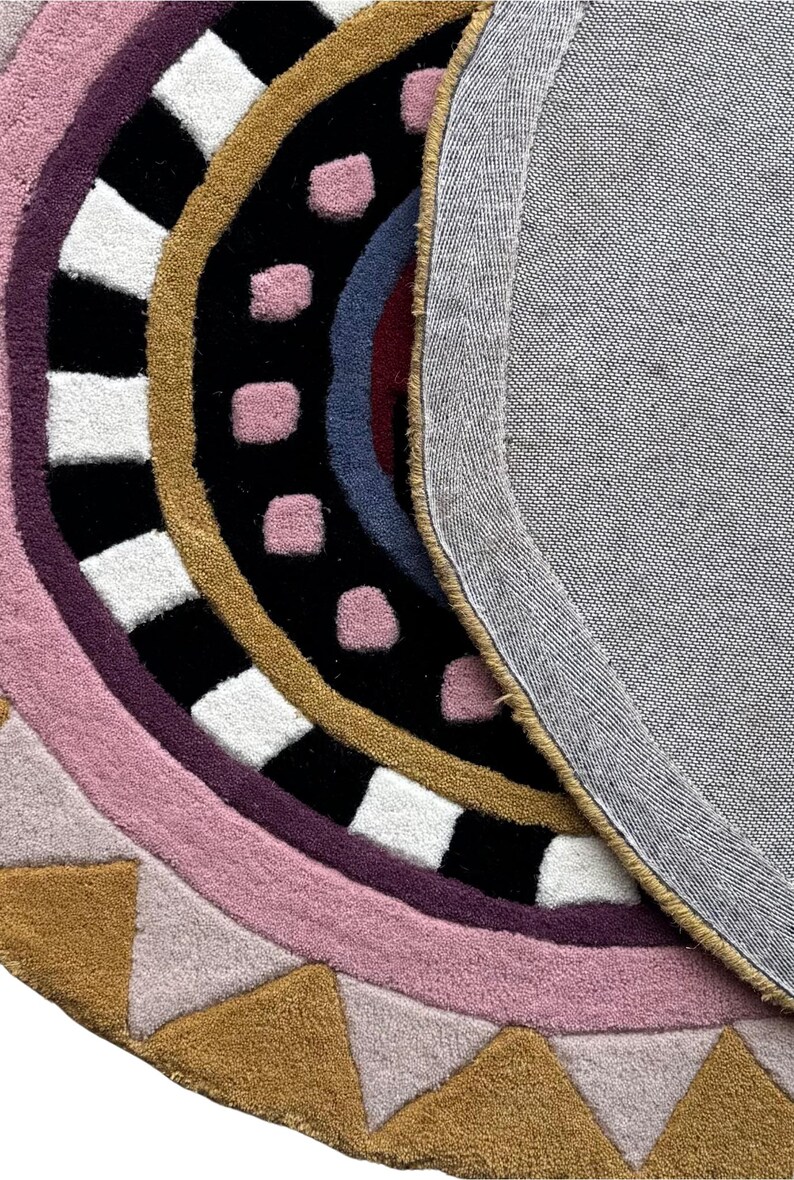 K&ouml;nnte beinhalten: Ein farbenfroher, runder Teppich mit geometrischem Design. Der Teppich hat konzentrische Farbstreifen, darunter Rosa, Lila, Schwarz, Gold und Wei&szlig;. Die R&uuml;ckseite ist ein grauer Stoff. Das Design umfasst Quadrate, Dreiecke und Punkte.