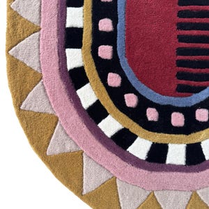 K&ouml;nnte beinhalten: Ein farbenfroher, halbkreisf&ouml;rmiger Teppich mit geometrischem Design. Der Teppich hat einen senfgelben Rand mit hellrosa Dreiecken, einen rosa Innenrand und ein schwarz-wei&szlig;es Schachbrettmuster. Die Mitte hat einen roten Bereich mit schwarzen Streifen.