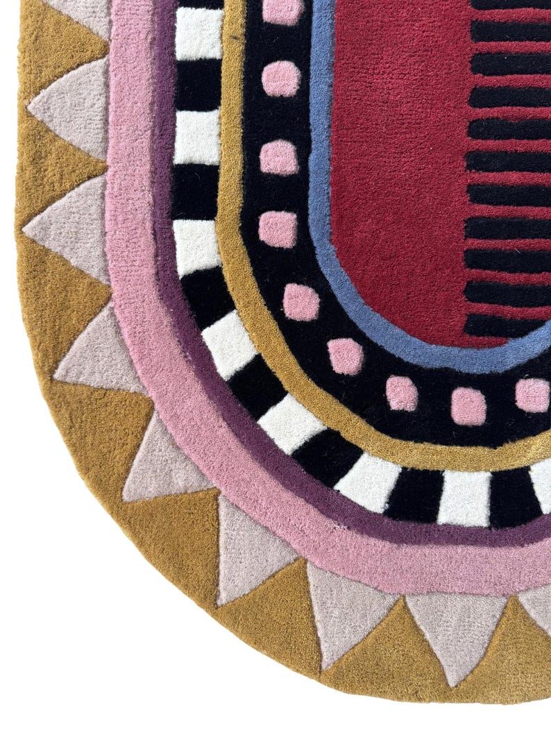 K&ouml;nnte beinhalten: Ein farbenfroher, halbkreisf&ouml;rmiger Teppich mit geometrischem Design. Der Teppich hat einen senfgelben Rand mit hellrosa Dreiecken, einen rosa Innenrand und ein schwarz-wei&szlig;es Schachbrettmuster. Die Mitte hat einen roten Bereich mit schwarzen Streifen.