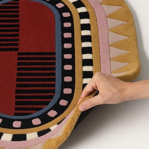 K&ouml;nnte beinhalten: Ein farbenfroher, ovaler Teppich mit geometrischem Design. Der Teppich hat ein rot-schwarzes Muster in der Mitte, umgeben von konzentrischen B&auml;ndern in Schwarz, Wei&szlig;, Rosa und Gold. Der Text "Pile height shown" ist unten zu sehen.