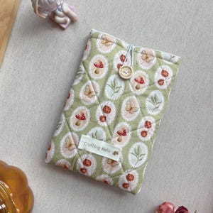 May include: A quilted green book sleeve with a repeating pattern of mushrooms, butterflies, and leaves in oval frames. The sleeve has a button closure and a label that reads "Crafting Kela."