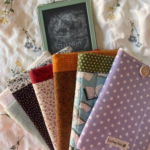 Könnte beinhalten: Eine Sammlung von sechs Stoffhüllen für E-Reader, die fächerförmig angeordnet sind. Die Hüllen zeigen verschiedene Muster wie Sterne, Blumenprints und offene Bücher. Eine hellviolette Hülle mit weißen Sternen hat einen Holzknopf und ein Etikett.