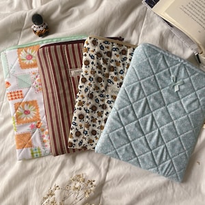 Könnte beinhalten: Vier rechteckige Stofftaschen mit Reißverschlüssen. Eine zeigt ein weißes, orangefarbenes und grünes Blumen-Patchwork-Muster. Eine andere hat vertikale rote und braune Streifen. Die dritte hat einen braunen und blauen Blumendruck. Die vierte ist hellblau gesteppt.