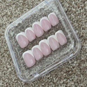 May include: A set of ten artificial nails in a clear plastic case. Each nail has a pale pink base with a white French tip. The nails are oval-shaped and have a glossy finish.