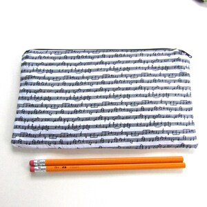 May include: A white zippered pencil case with a black and white musical note pattern. Two orange pencils are lying next to the case.