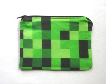 May include: A green and black pixelated zippered pouch. The pouch is made of fabric and has a zipper closure.