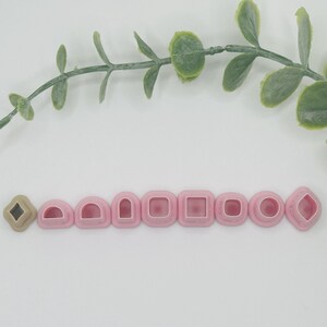 May include: A collection of pink polymer clay cutters in various geometric shapes, including a diamond, half-circle, square, and circle. A single tan diamond-shaped cutter is also present. A green leafy branch is in the background.
