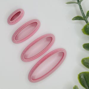 May include: Four pink oval-shaped cookie cutters of varying sizes are arranged on a white surface. The cutters have a raised edge and a hollow center. Green leafy sprigs are in the background.