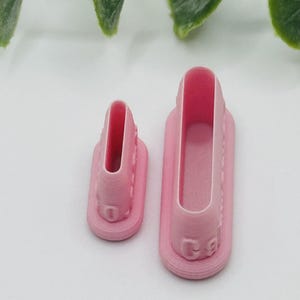 May include: Two pink plastic cookie cutters in the shape of a shoe. The cutters are different sizes, with a rectangular base and a shoe-shaped interior. The cutters have a ridged design around the base and interior.