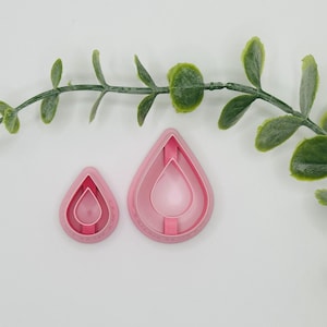 May include: Two pink teardrop-shaped cookie cutters, one large and one small, nested together. A green leafy branch curves across the top. The cutters have a layered design, perfect for baking and crafting projects.