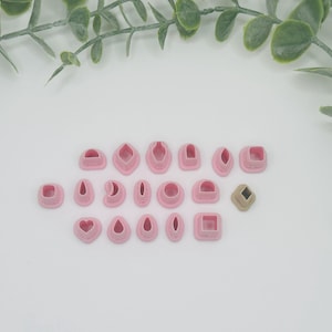 May include: A collection of pink polymer clay cutters in various shapes, including squares, hearts, and teardrops. One cutter is a tan diamond shape. The cutters are arranged on a white surface, with green leafy sprigs in the background.