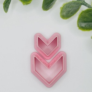 May include: Two pink chevron-shaped cookie cutters. The cutters are nested, with a smaller one on top of a larger one. The edges are a darker pink than the centers. The background is white with green leaves.