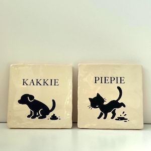 May include: Two off-white square ceramic tiles with black illustrations. One tile shows a dog with the word "KAKKIE", the other shows a cat with the word "PIEPIE". The dog and cat are depicted in a playful, cartoon style.