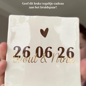 May include: A square white ceramic tile with a brown heart and gold lettering. The tile features the date "26.06.26" and the names "Wout & Nina". The text above reads "Het trouwseizoen is weer begonnen!" and "Geef dit leuke tegeltje cadeau aan het bruidspaar!"